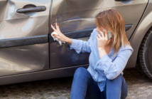 Blonde female in casual attire sitting near dented car and touching scratches and dents on automobile door, talking phone with insurance agent or ask help, road accident. Outdoor shot.