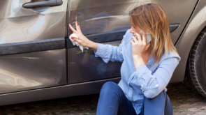 Blonde female in casual attire sitting near dented car and touching scratches and dents on automobile door, talking phone with insurance agent or ask help, road accident. Outdoor shot.
