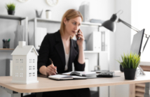 Beautiful young girl standing in a bright office near the desktop. The girl has blond hair. She is wearing a light blouse and a striped suit. photo with depth of field