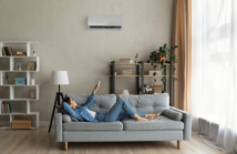 Glad millennial female buyer of air conditioner relax on sofa hold controller breath cool fresh air despite of hot summer day outside. Happy young lady regulate climate at home using modern ac device