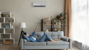 Glad millennial female buyer of air conditioner relax on sofa hold controller breath cool fresh air despite of hot summer day outside. Happy young lady regulate climate at home using modern ac device