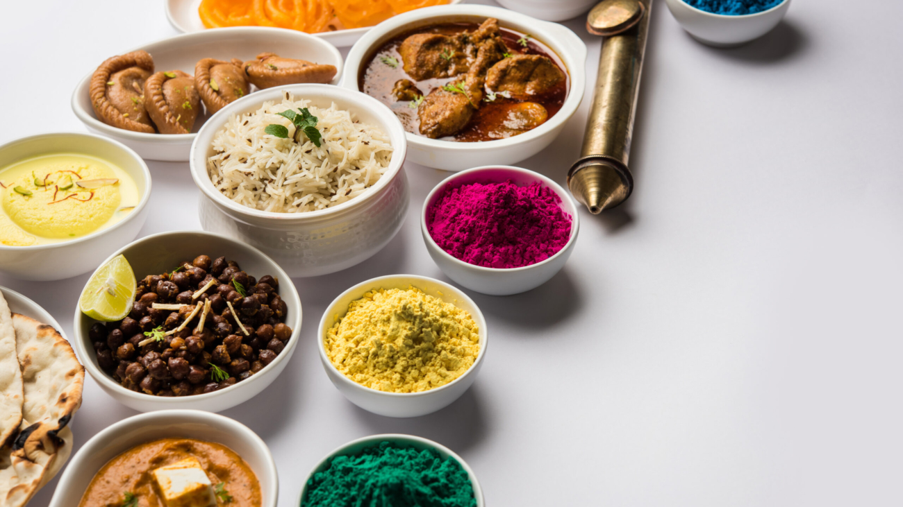 Happy Holy concept showing Indian assorted lunch food like chicken, paneer butter masala, naan, jeera rice, black chana fry, jalebi, ras malai, thandai and Farsan with holi colours and pichkari